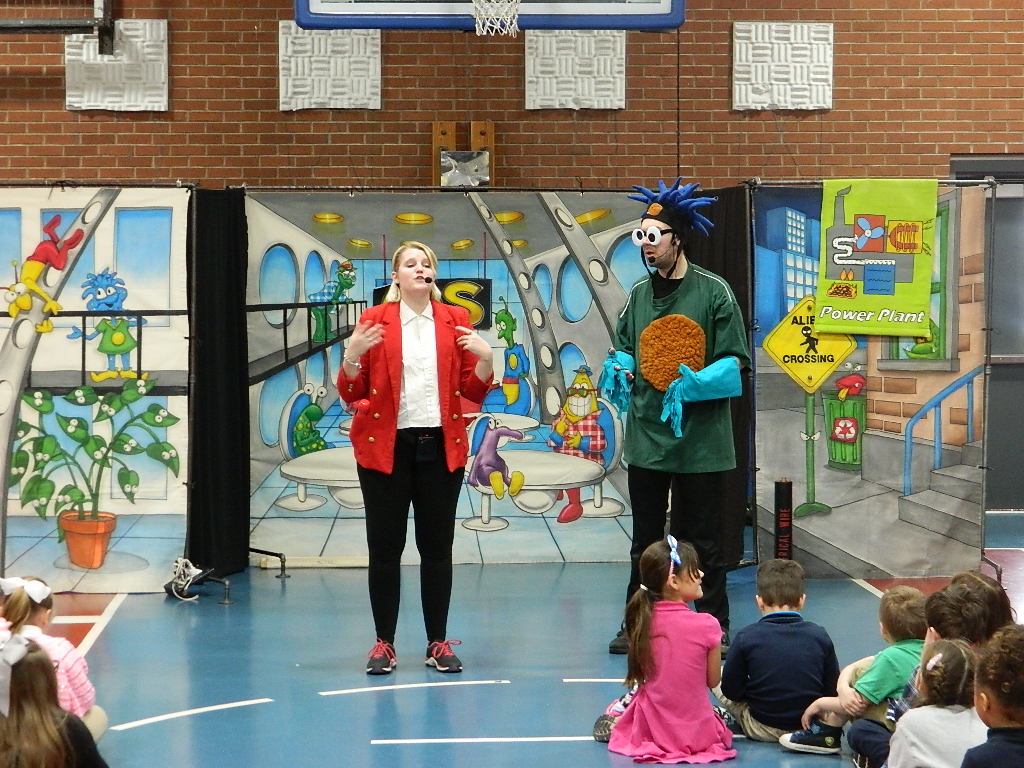 Woman in red blazer, man in green monster suit talking to children on an elementary school floor