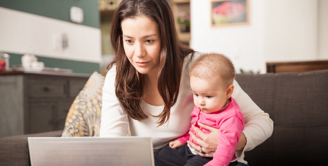 Mom sitting on sofa with baby on lap, looking at laptop screen