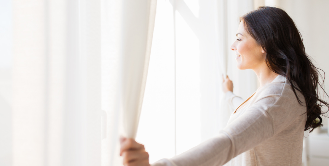 Woman opening window curtains to let in bright sunlight