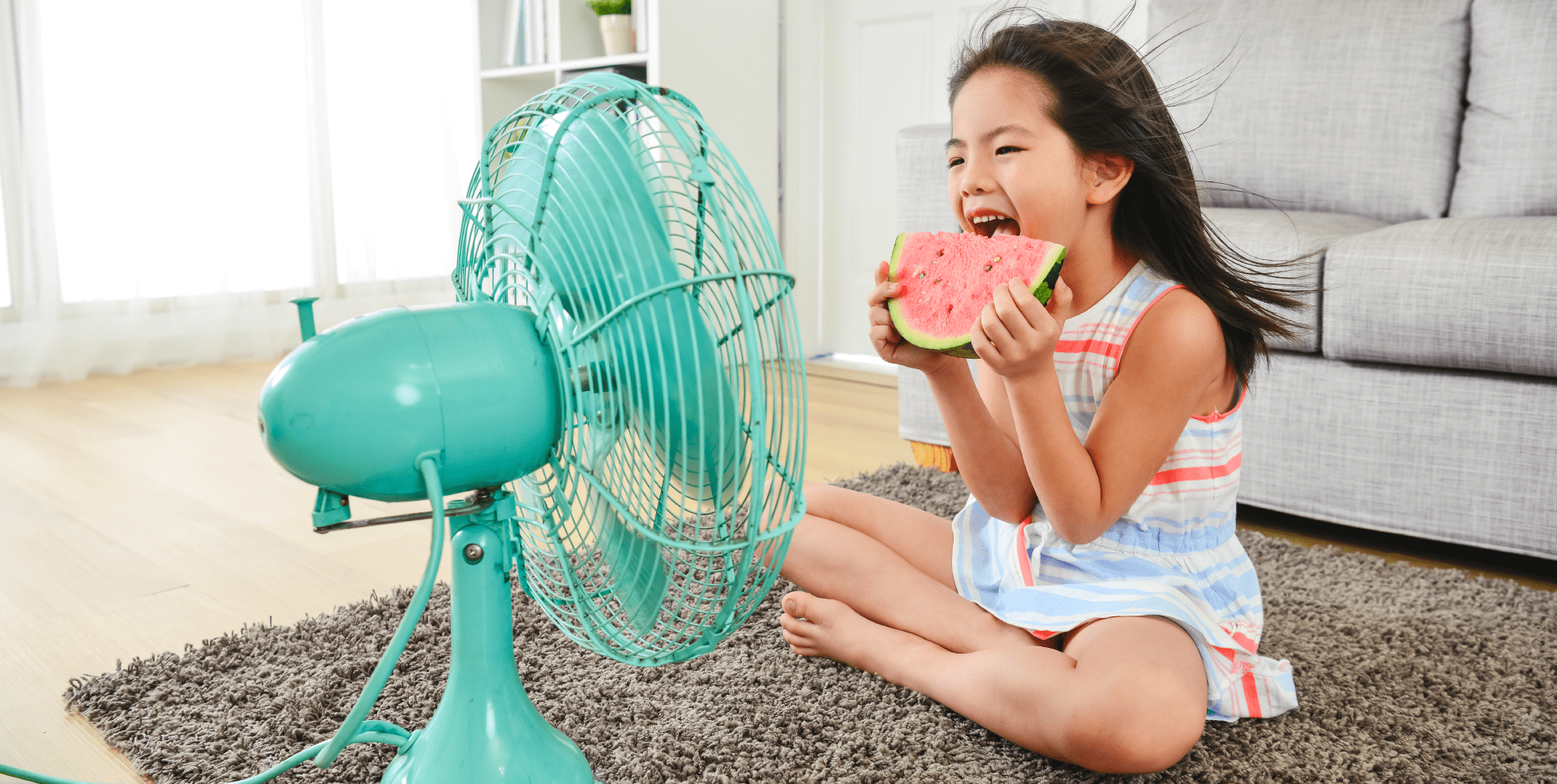 Child sitting on the floor facing a portable room fan and eating a slice of watermelon