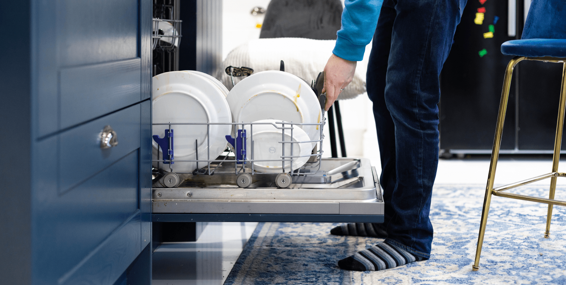 A person’s legs are visible as they load dirty dishes into the bottom rack of a dishwasher. 
