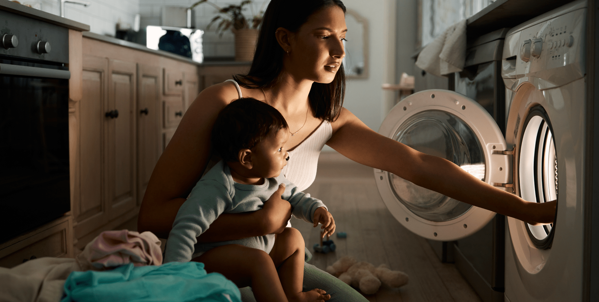 A woman holding a baby kneels in front of a clothes dryer in dim lighting with the light from the dryer reflected on her face. 