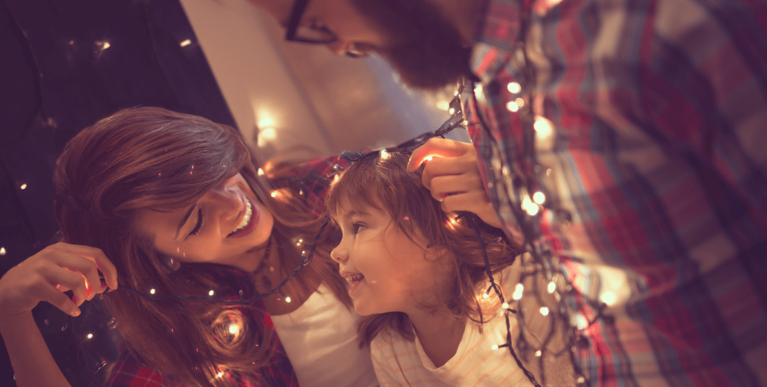 Two parents and a child smiling at each other while holding a string of holiday lights.