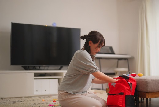 A woman is kneeling on the floor, carefully packing a bright red emergency backpack. The scene appears to be indoors, possibly in a living room, with a soft carpet and a tablet placed nearby on the wooden floor. The room is well-lit, and there are toys scattered around, indicating a family environment. The woman seems focused on organizing the contents of the backpack, suggesting preparedness or planning for an emergency situation.