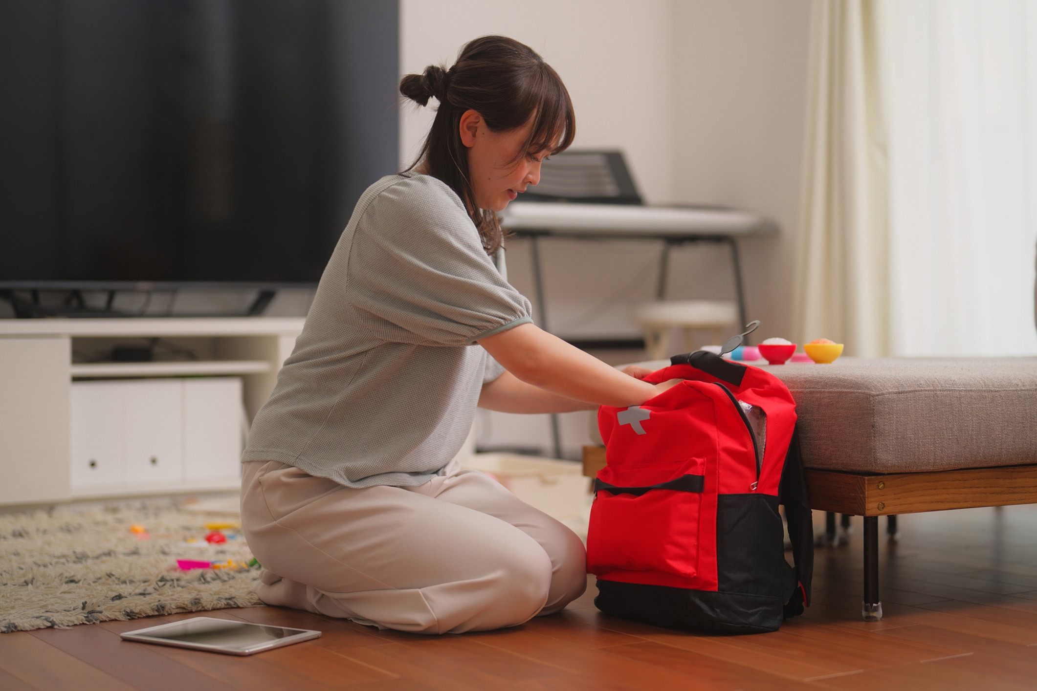 A woman is kneeling on the floor, carefully packing a bright red emergency backpack. The scene appears to be indoors, possibly in a living room, with a soft carpet and a tablet placed nearby on the wooden floor. The room is well-lit, and there are toys scattered around, indicating a family environment. The woman seems focused on organizing the contents of the backpack, suggesting preparedness or planning for an emergency situation.