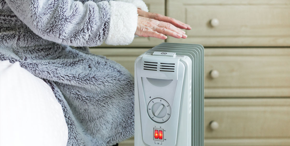 Elderly person warming hands over a space heater in a cozy room.