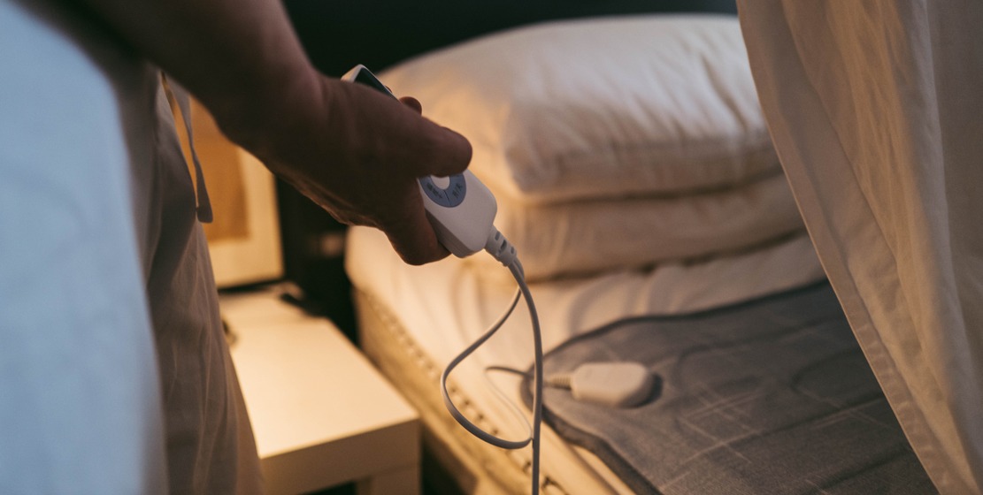 Hand reaching for an electric heating pad controller on a bed with white bedding in dim, warm lighting