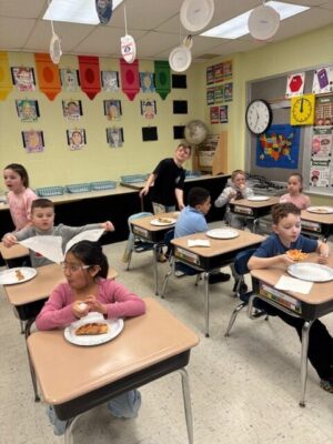 An elementary classroom where several children sit at individual desks eating slices of pizza on paper plates. The room has colorful wall decorations, student artwork, a clock, and hanging paper shapes, with desks arranged in rows.