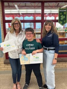 Three people stand in a school hallway in front of red-framed windows and a library area. The person in the center holds a colorful drawing and a certificate labeled "Ideas." The two people beside the student also hold matching certificates.