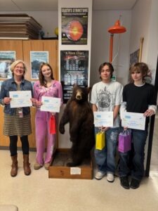 Four people stand in a classroom holding certificates and small gift bags. A mounted black bear specimen stands on a wooden base in the center of the group. The room contains science posters on the wall, including charts labeled "Earth's Structure" and "A Rock Chart," with shelves and classroom materials in the background.