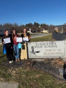 Three people stand outdoors beside a stone sign that reads "Susquenita High School, Home of the Blackhawks." Each person holds a certificate, and one also holds a small gift bag. The scene shows the school grounds with grass, a sidewalk, and buildings in the background under a clear blue sky.