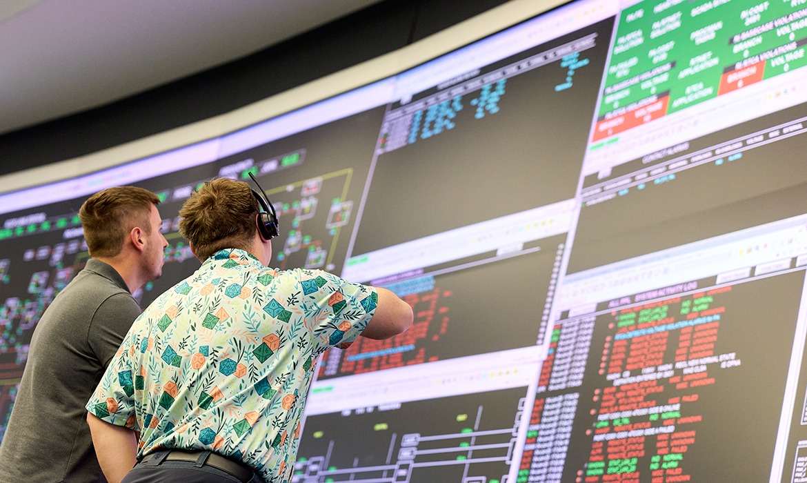 Two men stand in front of numerous screens with data and information.