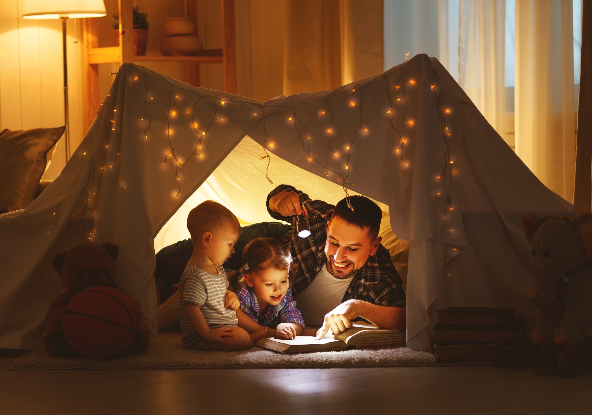 Father reading a book by flashlight with two young children inside a blanket fort lit by string lights